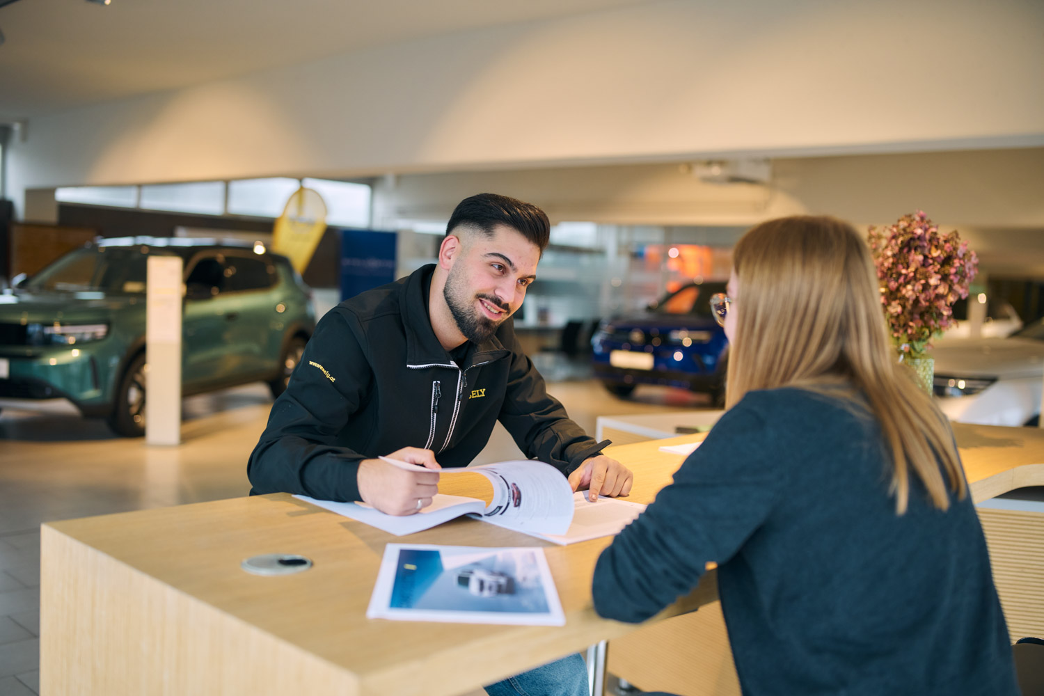 Verkäufer und Kundin sitzen an einem Tisch im Autohaus und blättern gemeinsam durch ein Prospekt.